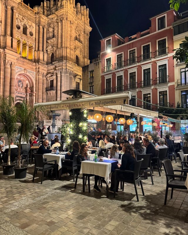 Een vol terras bij de verlichte kathedraal van Málaga in de avond