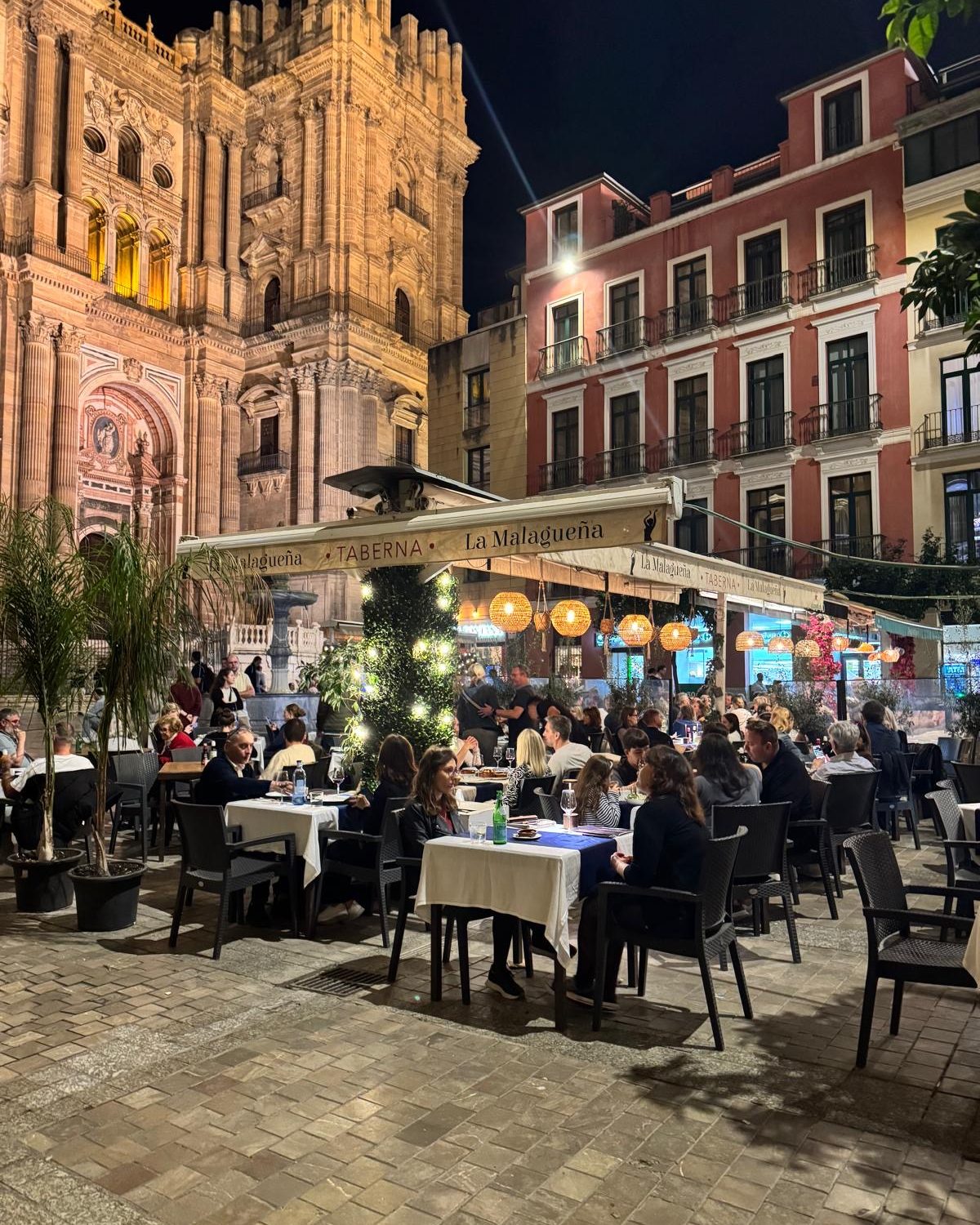 Een vol terras bij de verlichte kathedraal van Málaga in de avond